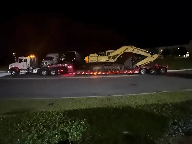 Yellow excavator chained on a red lowboy trailer at night behind a white Kenworth tractor